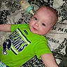 baby, child, infant, smile, seahawks, green_shirt, bottle, bedspread, pattern, blue_eyes, cute, lying_down, plaid_pants, person, indoor, soft_lighting, happy, face, head, clothing