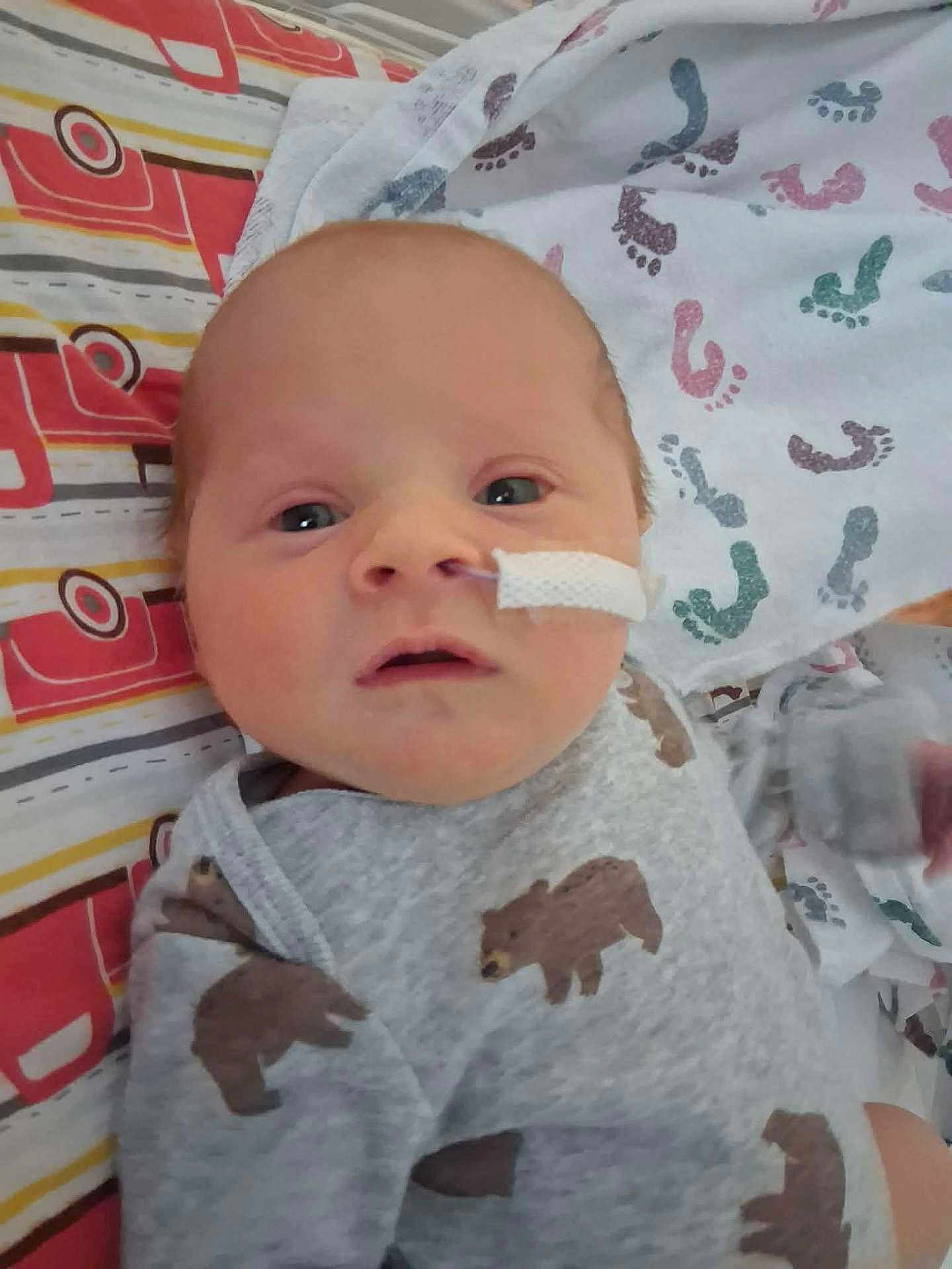 John is registered to the contest to win money with this photo: baby, newborn, infant, onesie, bear_print, medical_tape, nasal_tube, bed, blanket, footprint_pattern, face, head, person, clothing, cute, lying_down, indoor, closeup, soft_lighting, expression