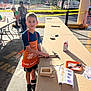 Daniel joined the competition — help win amazing prizes! child, boy, apron, hammer, workshop, table, outdoor, daylight, caution_tape, parking_lot, tools, wooden_box, instruction_sheet, person, shoes, shorts, trees, sign, car, event