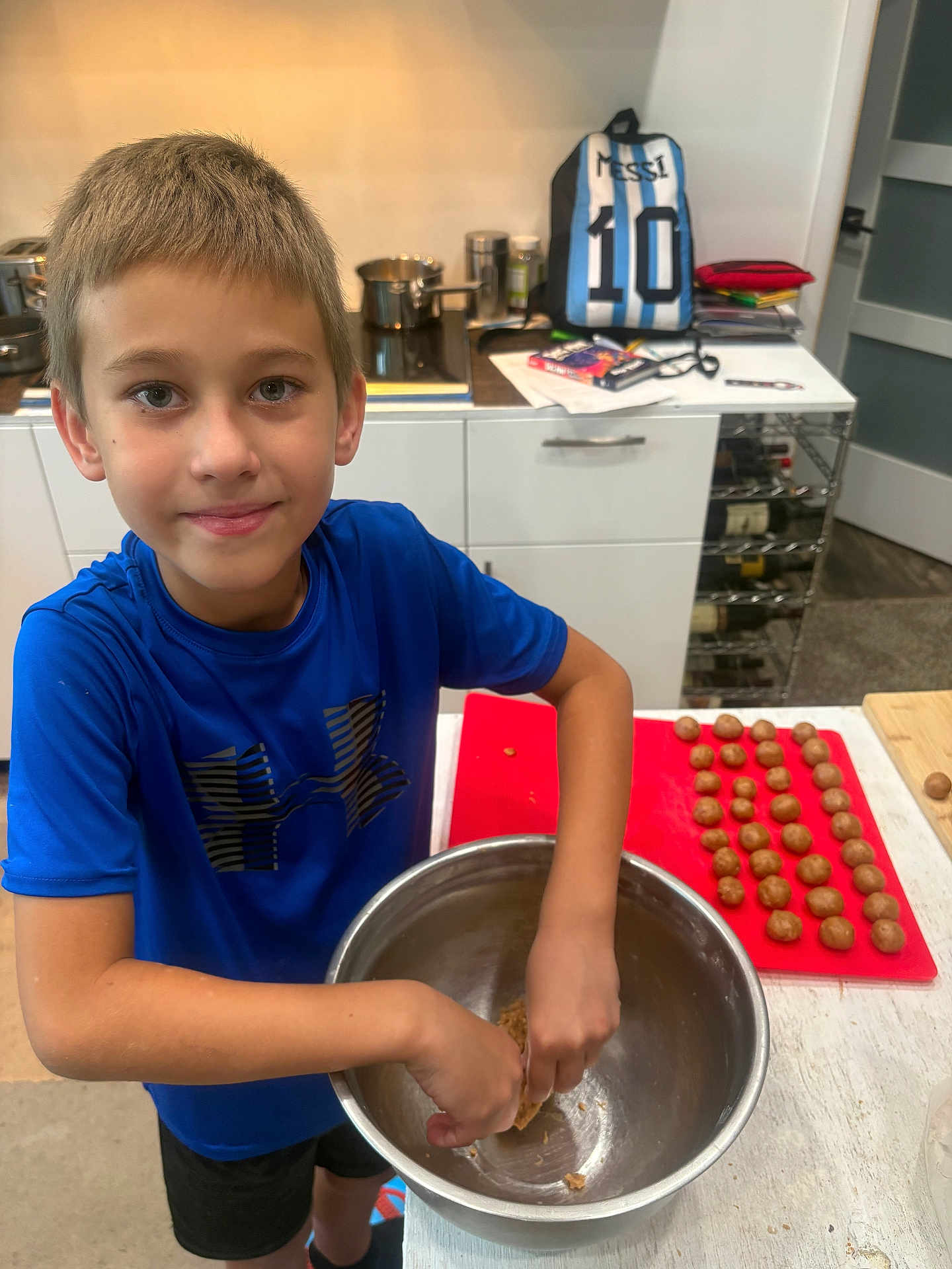 Daniel joined the competition — help win amazing prizes! boy, child, blue_shirt, kitchen, mixing_bowl, dough, cutting_board, rolled_dough, backpack, messy_hair, smiling, cooking, indoors, countertop, pots, stove, cabinet, person, shorts, preparing_food