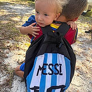 Mark is registered to the contest to win money with this photo: child, hug, backpack, messy_hair, outdoor, sunlight, grass, leaves, car, trailer, boy, blue_shirt, red_shirt, hand, nature, casual, portrait, siblings, affection, toddler