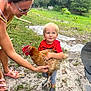 child, toddler, woman, chicken, animal, grass, outdoor, nature, sand, dirt, holding, smiling, red_shirt, glasses, footwear, greenery, daylight, person, interaction, playful