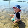 toddler, child, water, lake, hat, bucket_hat, summer, sunny, outdoor, nature, ripples, swim_shorts, sitting, reflection, clear_water, sky, shore, daytime, young_child, casual_clothing