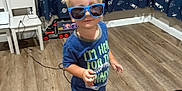 Mark is registered to the contest to win money with this photo: child, toddler, sunglasses, blue_shirt, printed_pants, barefoot, wooden_floor, indoor, toy_truck, chairs, curtains, space_pattern, blond_hair, standing, playful, smiling, person, room, cord, casual_clothing
