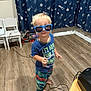 child, toddler, sunglasses, blue_shirt, printed_pants, barefoot, wooden_floor, indoor, toy_truck, chairs, curtains, space_pattern, blond_hair, standing, playful, smiling, person, room, cord, casual_clothing