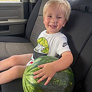 Mark is registered to the contest to win money with this photo: child, smiling, watermelon, car_seat, seatbelt, blond_hair, tshirt, nike_logo, window, outdoor_view, happy, portrait, summer, fruit, hand, casual, interior, seat, person, young