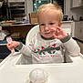 Mark is registered to the contest to win money with this photo: toddler, child, high_chair, fork, baseball, toy, kitchen, indoor, white_clothing, blond_hair, facial_expression, baby, table, furniture, plastic_utensil, carpet, cabinet, appliance, person, cute