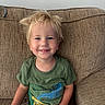 child, smile, blond_hair, green_shirt, dinosaur_print, sippy_cup, couch, brown_couch, indoor, happy, casual, toddler, portrait, seated, young_child, comfortable, home, relaxed, person, cute