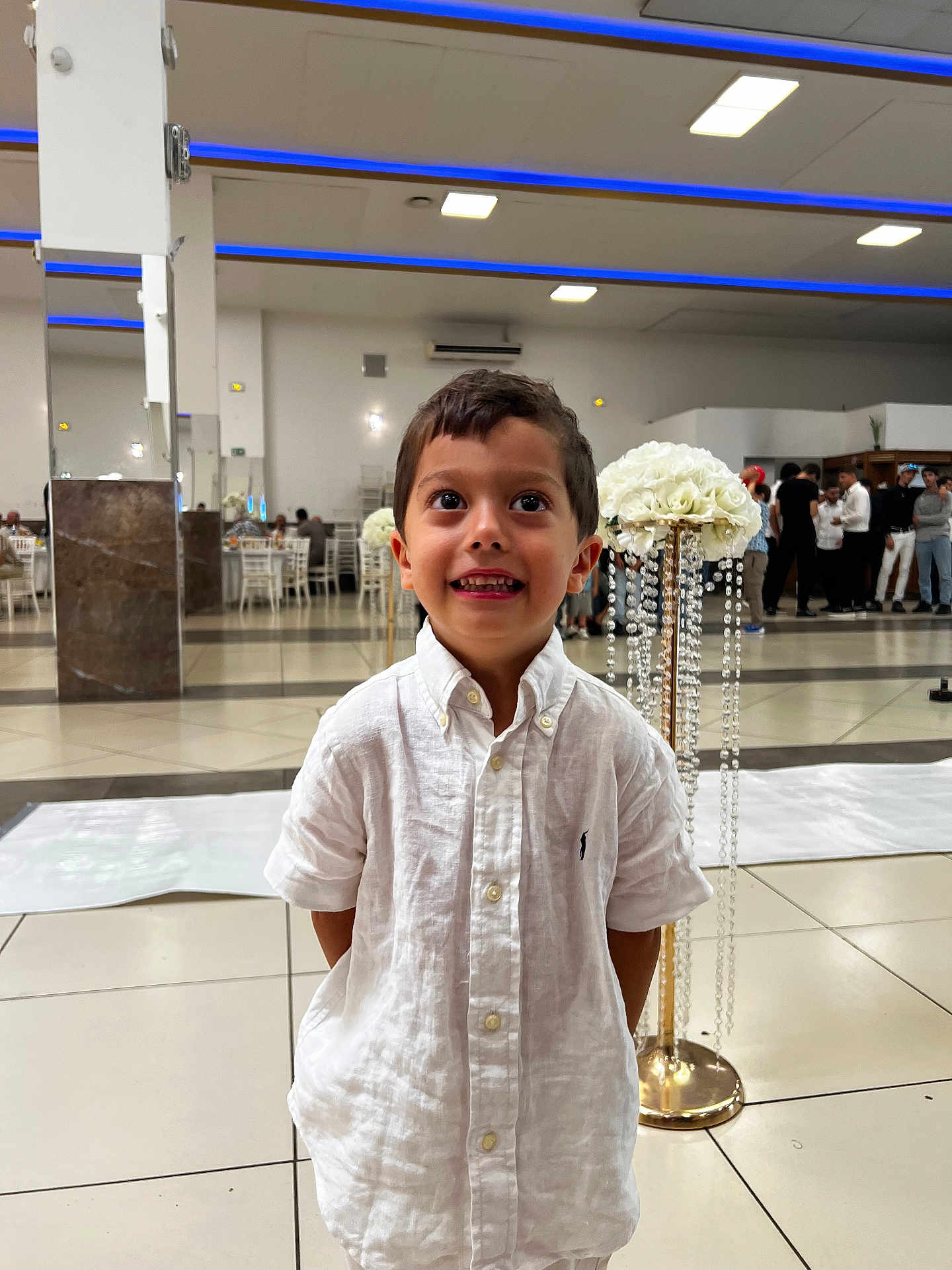 Imran participe au concours pour gagner de l'argent avec cette photo : child, boy, white_shirt, smile, indoor, event, flower_arrangement, people, formal, floor_tiles, group, celebration, decor, happy, portrait, standing, hands_behind_back, short_hair, casual, lighting