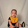 baby, child, infant, sitting, yellow_bib, black_clothing, sneakers, footwear, portrait, studio, neutral_background, curious_expression, young_child, indoors, cute, person, casual, fashion, accessory, chain_necklace