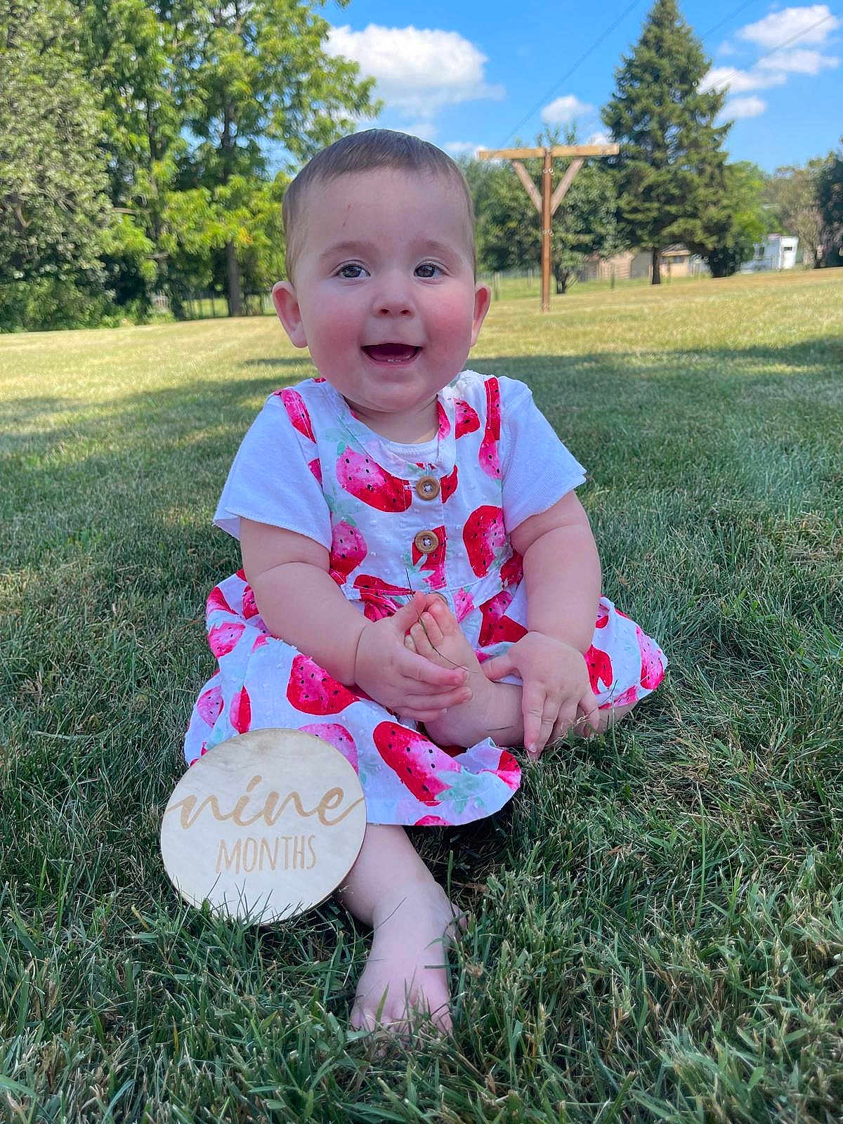 Oaklynn is registered to the contest to win money with this photo: baby, child, grass, outdoor, smiling, dress, watermelon_print, foot, hand, sign, nine_months, sunny, blue_sky, tree, park, nature, happy, infant, sitting, cute