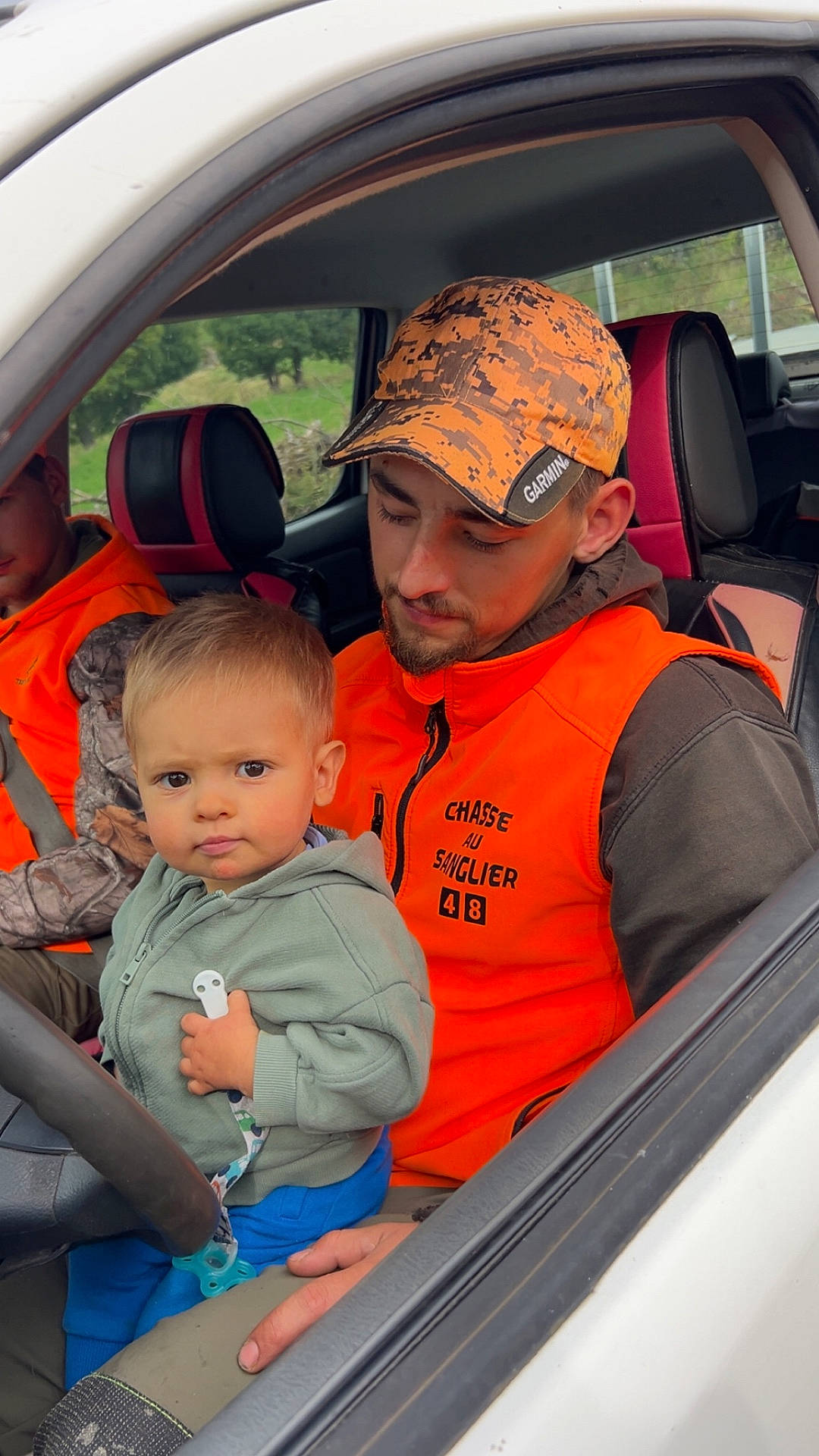 Nino a rejoint le concours — aidez-le/la à gagner de superbes lots ! camouflage, cap, car_interior, casual_clothing, child, driving_wheel, greenery, holding, looking_down, man, nature, orange_vest, outdoor, pacifier, person, seat, seatbelt, toddler, vehicle, window
