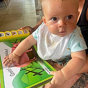 Leo participe au concours pour gagner de l'argent avec cette photo : arm, baby, bib, book, child, clothing, curious, expression, floor, hand, holding, indoor, light, person, pig, portrait, reading, tattoo, tile, toy