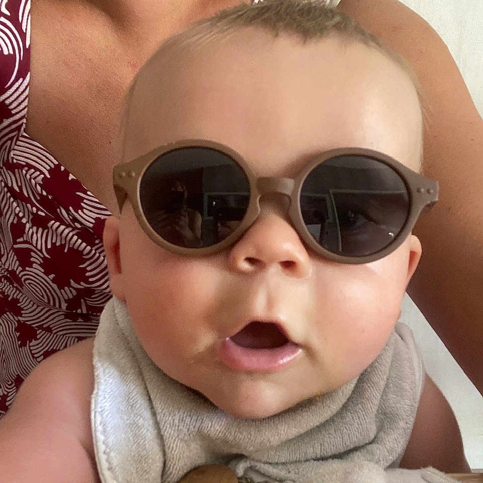 Leo participe au concours pour gagner de l'argent avec cette photo : accessory, adult, arm, baby, beads, bib, casual, child, closeup, expression, face, head, indoor, mouth_open, pacifier_clip, patterned_clothing, person, skin, sunglasses, wooden_surface