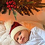 baby, infant, christmas_hat, red_hat, white_hat, blanket, christmas_tree, pine_branches, christmas_lights, red_ornaments, holiday, festive, cozy, indoors, cute, child, portrait, sleepwear, soft_texture, holiday_decor