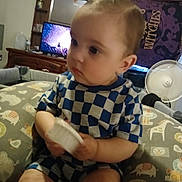 Kash joined the competition — help win amazing prizes! toddler, child, baby, blue_checkered_clothing, cushion, animal_print, indoor, fan, television, tapestry, wooden_furniture, curious, holding_object, seated, face, head, short_hair, room, patterned_fabric, home