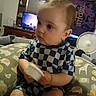 toddler, child, baby, blue_checkered_clothing, cushion, animal_print, indoor, fan, television, tapestry, wooden_furniture, curious, holding_object, seated, face, head, short_hair, room, patterned_fabric, home