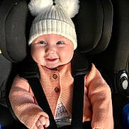 Rosie is registered to the contest to win money with this photo: baby, infant, car_seat, knit_hat, pom_poms, cardigan, smiling, happy, child, seatbelt, cute, face, clothing, indoor, person, warm_clothes, portrait, baby_hats, closeup, child_seat