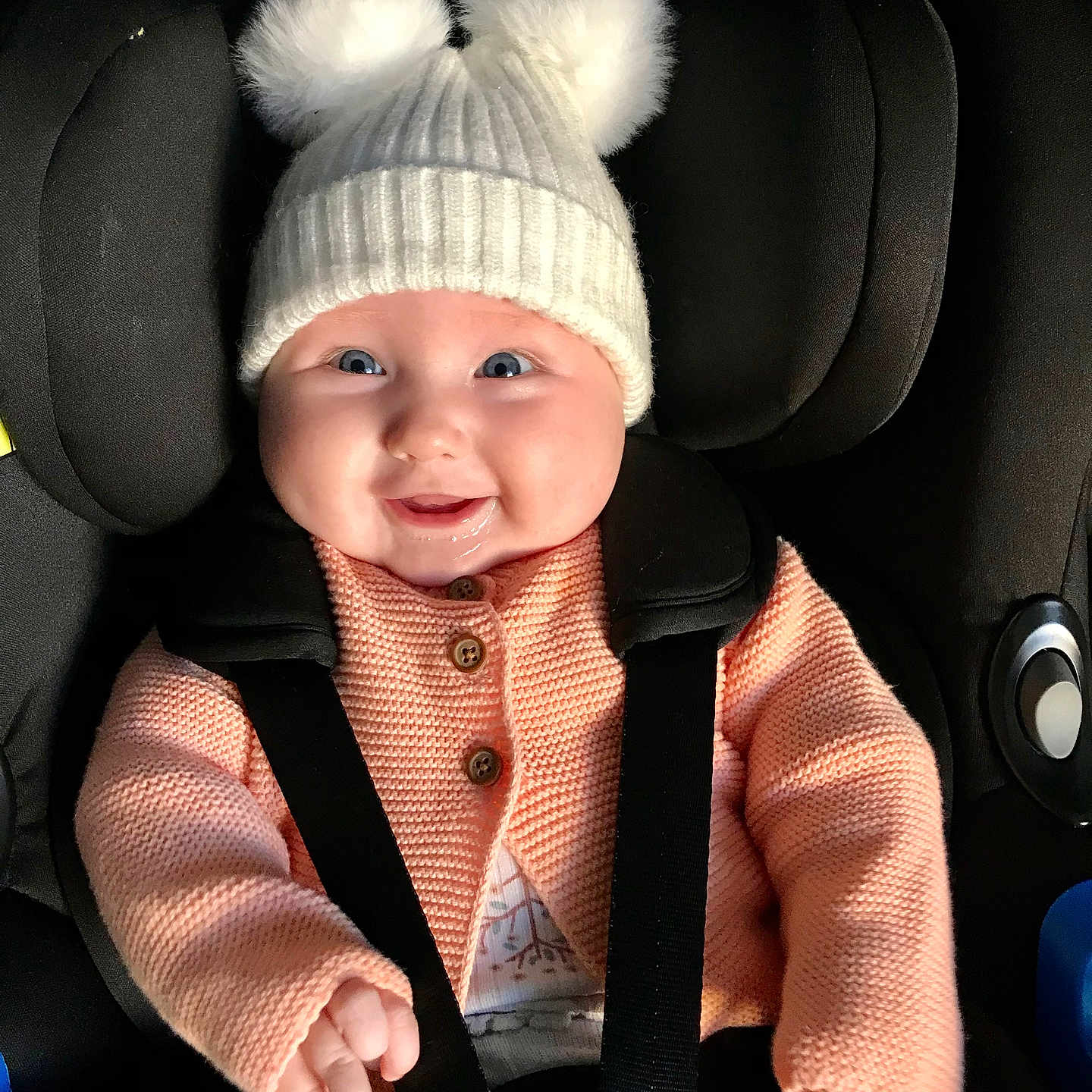 Rosie is registered to the contest to win money with this photo: baby, baby_hats, car_seat, cardigan, child, child_seat, closeup, clothing, cute, face, happy, indoor, infant, knit_hat, person, pom_poms, portrait, seatbelt, smiling, warm_clothes