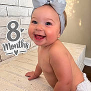 Oaklyn joined the competition — help win amazing prizes! baby, infant, child, smiling, happy, portrait, sitting, bow, headband, diaper, milestone, eight_months, indoor, mattress, bedroom, brick_wall, warm_lighting, photography, text_overlay, closeup
