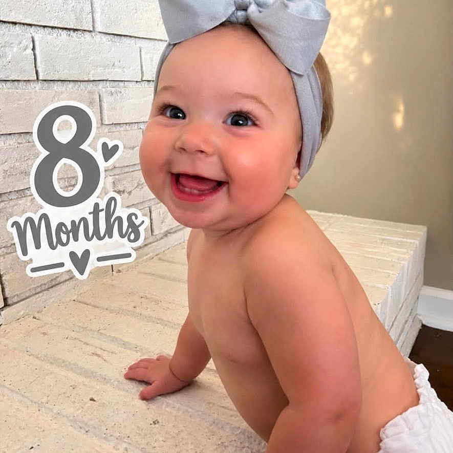 Oaklyn joined the competition — help win amazing prizes! baby, infant, child, smiling, happy, portrait, sitting, bow, headband, diaper, milestone, eight_months, indoor, mattress, bedroom, brick_wall, warm_lighting, photography, text_overlay, closeup