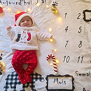 Amélia participe au concours pour gagner de l'argent avec cette photo : baby, infant, christmas, holiday, santa_hat, red_pants, white_sweater, lying_down, blanket, calendar, numbers, decorations, string_lights, candy_cane, paw_prints, smiling, cute, festive, indoors, portrait