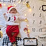 baby, infant, christmas, holiday, santa_hat, red_pants, white_sweater, lying_down, blanket, calendar, numbers, decorations, string_lights, candy_cane, paw_prints, smiling, cute, festive, indoors, portrait