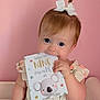 baby, bow, chewing, child, cute, dress, eyes, face, hair, hand, indoors, infant, milestone_card, person, pink_background, portrait, sitting, skin, smile, strawberry_print