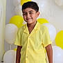 child, boy, yellow_clothing, smile, balloons, party, celebration, yellow_balloon, white_balloon, flower_balloon, shorts, buttoned_shirt, standing, happy, indoor, portrait, festive, decorations, cute, casual