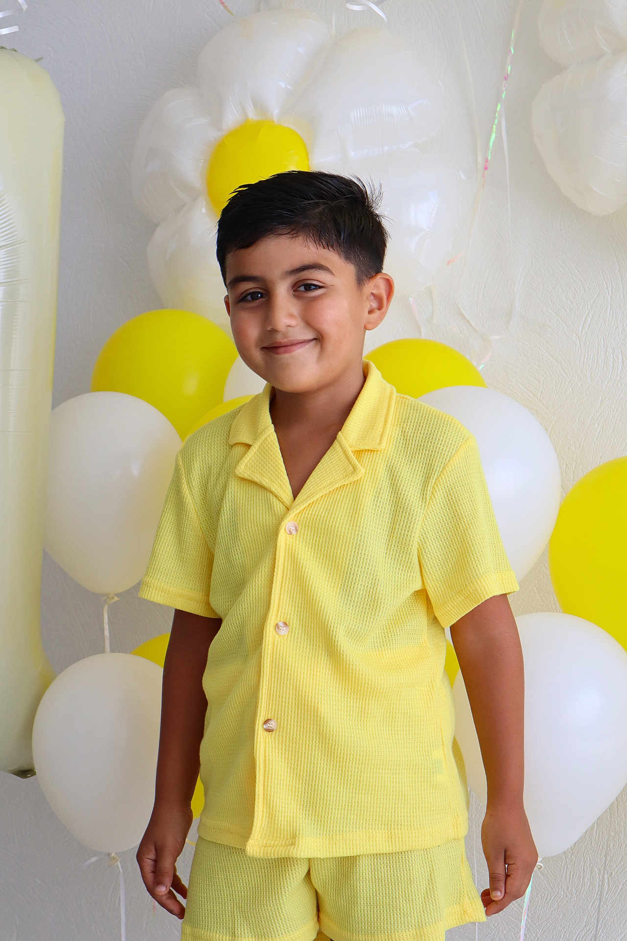 Eymen Asaf participe au concours pour gagner de l'argent avec cette photo : child, boy, yellow_clothing, smile, balloons, party, celebration, yellow_balloon, white_balloon, flower_balloon, shorts, buttoned_shirt, standing, happy, indoor, portrait, festive, decorations, cute, casual