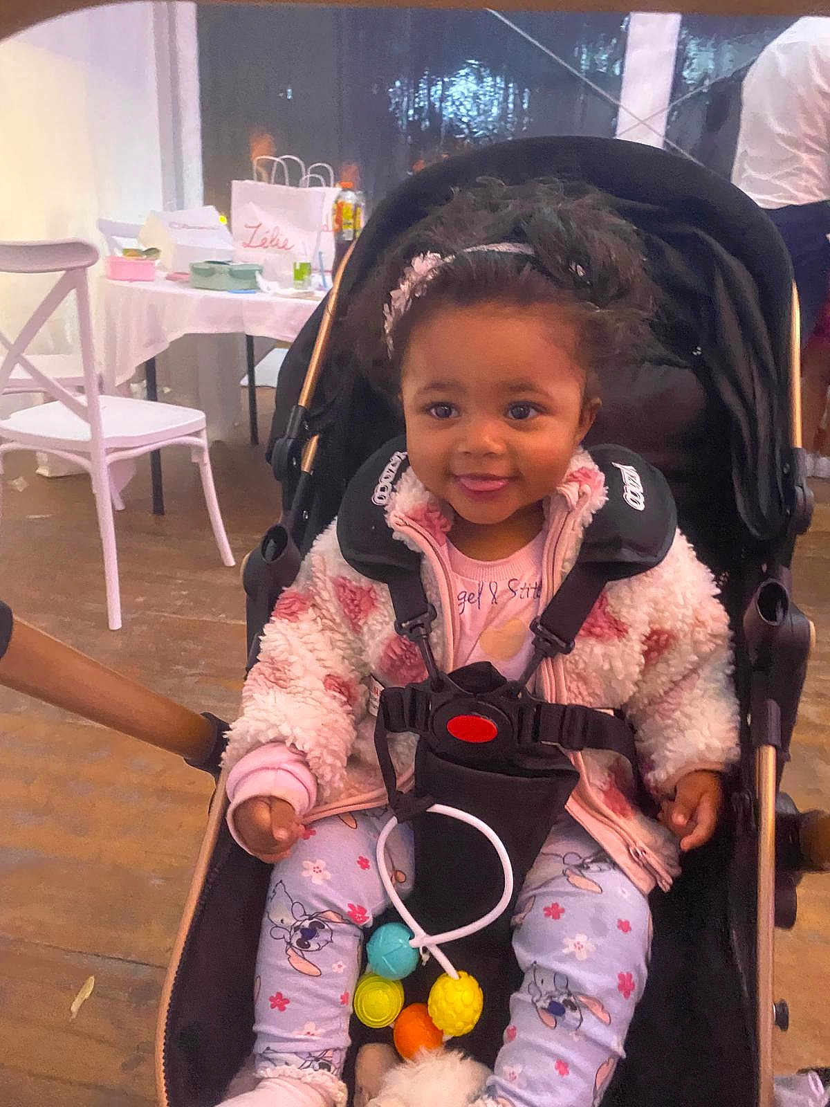 Manon participe au concours pour gagner de l'argent avec cette photo : toddler, child, stroller, jacket, pajamas, headband, curly_hair, indoors, table, chair, toy, smiling, cute, baby, playful, seated, person, clothing, floor, casual