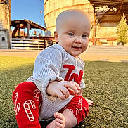 Rayah joined the competition — help win amazing prizes! baby, child, infant, sitting, grass, outdoor, sunlight, blue_eyes, red_pants, candy_cane_pattern, beige_sweater, smiling, cute, barefoot, portrait, daylight, industrial_background, curious, young, person