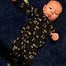 baby, infant, child, portrait, lying_down, blue_rug, black_outfit, printed_clothing, hey_text, waving, fist, bald, eyes_open, curious, indoors, soft_surface, full_body, casual, photograph, cute