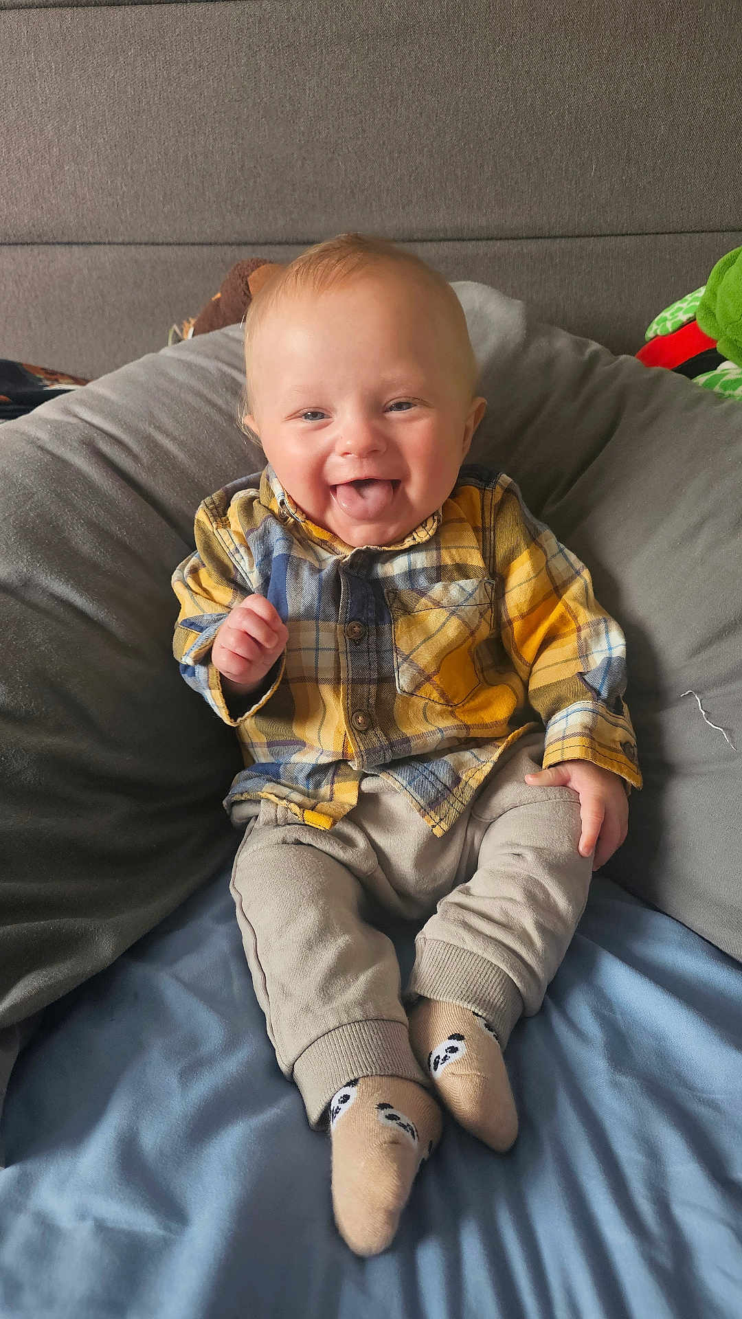 Leo joined the competition — help win amazing prizes! baby, child, smiling, tongue_out, plaid_shirt, panda_socks, pants, sitting, bed, pillow, cozy, happy, cute, indoors, soft_furniture, infant, clothing, person, portrait, casual
