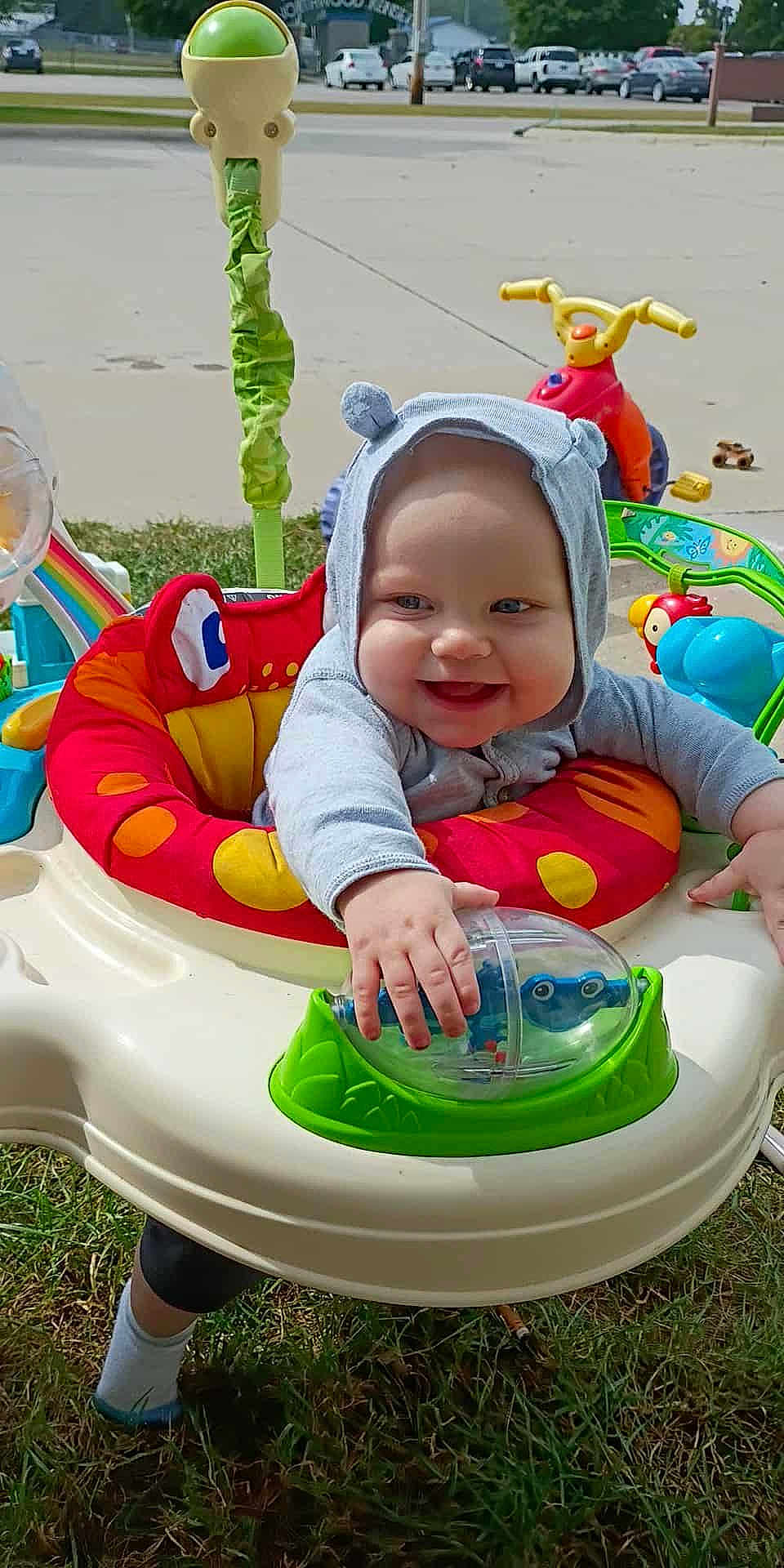 Wyatt is registered to the contest to win money with this photo: baby, child, smiling, hooded_clothing, activity_center, toy, outdoor, grass, playtime, colorful, happy, infant, cute, person, sitting, sunny, daytime, hand, footwear, play