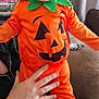 Wyatt joined the competition — help win amazing prizes! baby, child, pumpkin_costume, orange_clothing, smiling, happy, indoor, hand, socks, face, person, costume, holiday, halloween, living_room, bookshelf, curtain, couch, adult_hand, footwear