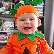 Wyatt joined the competition — help win amazing prizes! baby, child, costume, pumpkin_costume, orange_clothing, green_leaves, smiling, happy, face, headwear, indoor, bookshelf, dvds, shelf, person, cute, holiday, festive, portrait, young_child