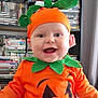 baby, child, costume, pumpkin_costume, orange_clothing, green_leaves, smiling, happy, face, headwear, indoor, bookshelf, dvds, shelf, person, cute, holiday, festive, portrait, young_child