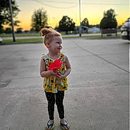 Adalynn is registered to the contest to win money with this photo: child, girl, flower, sunset, outdoor, concrete, polka_dot, leggings, flip_flops, yellow_top, hair_bun, smile, nature, trees, road, bench, car, evening, holding, cute