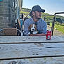 beagle, cap, casual, coast, companionship, daytime, dog, green_hills, hoodie, man, outdoor, people, picnic_table, puppy, railing, relaxed, seated, soda_can, sunny, wood