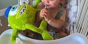 Chester is registered to the contest to win money with this photo: baby, child, high_chair, plush_toy, green_toy, sippy_cup, snacks, clapping, happy, indoor, feeding, seat, baby_food, toy, smiling, cute, person, infant, baby_clothing, home
