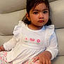toddler, child, girl, sitting, couch, white_top, floral_pants, indoors, casual_clothing, curly_hair, brown_eyes, portrait, cute, young_child, comfortable, relaxed, home, expression, fashion, furniture
