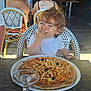 casual, chair, cheese, child, curly_hair, daylight, food, glass, happy, indoor, olive, person, pizza, plate, restaurant, seat, smiling, table, toddler, white_shirt