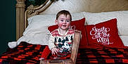 Ariana joined the competition — help win amazing prizes! bed, blanket, book, child, christmas_decor, face, festive, holiday, indoors, mickey_mouse, person, plaid, reading, red_clothing, red_pillow, smiling, sweater, toddler, white_pillow, wooden_headboard