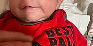Elijah is registered to the contest to win money with this photo: baby, infant, newborn, close_up, face, eyes, nose, mouth, skin, red_shirt, clothing, text_on_shirt, pillow, bed, hand, caregiver_hand, portrait, curious, indoors, smiling