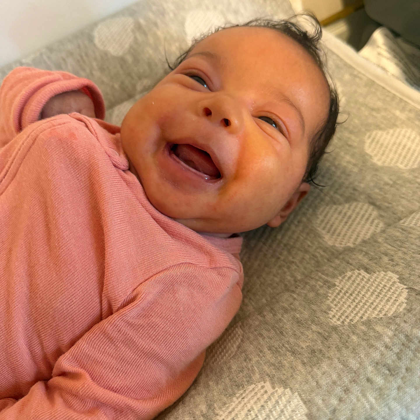 Zuri is registered to the contest to win money with this photo: baby, smiling, infant, pink_clothing, cushion, heart_pattern, lying_down, happy, portrait, face, child, cute, indoors, soft_texture, head, closeup, person, cozy, newborn, expression
