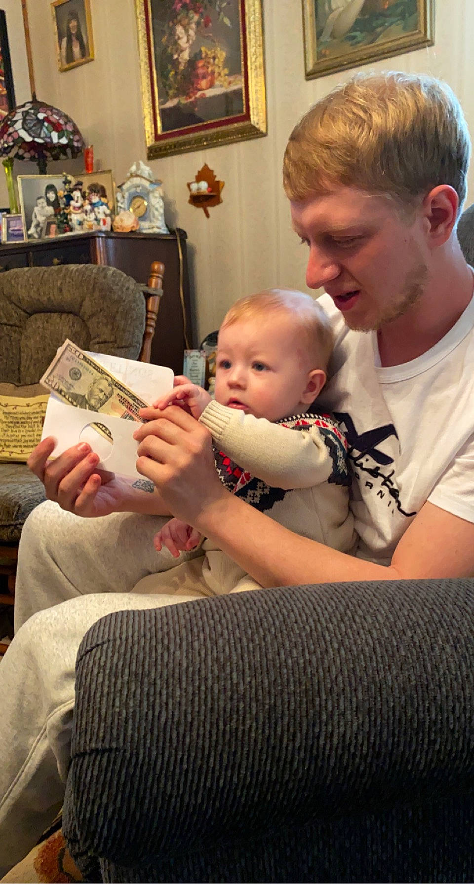 Rennen is registered to the contest to win money with this photo: baby, book, child, comfort, event, family, fun, happy, holiday, human, interaction, lamp, lap, living_room, person, picture_frame, room, shelf, sitting, t_shirt