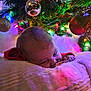 baby, blanket, christmas_tree, christmas_lights, ornament, decorations, festive, holiday, toddler, face, indoor, cozy, colorful, soft_texture, child, person, warm_lighting, greenery, red_clothing, reflection