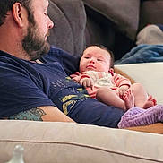 Eve is registered to the contest to win money with this photo: baby, baby_bottle, beard, blanket, clothing, couch, footwear, home, indoor, infant, man, pacifier, person, pillow, plant, portrait, relaxing, remote_control, sofa, tattoo