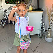 Aria participe au concours pour gagner de l'argent avec cette photo : toddler, child, indoor, floor_tiles, pigtails, white_top, green_shorts, black_shoes, pop_it_purse, butterfly_shaped, pink_container, curious_expression, living_room, tv_screen, high_chair, plant_decoration, glass_vase, mismatched_laces, socks, modern_interior